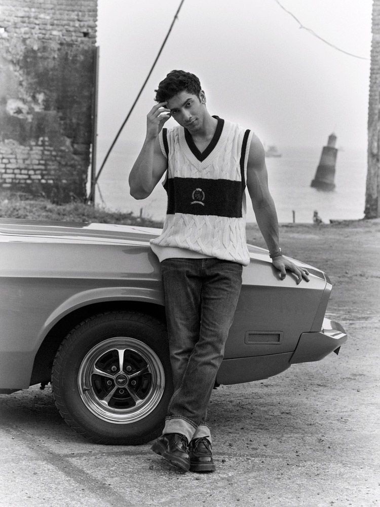 Vedang Raina leans against a car wearing a sweater vest and jeans