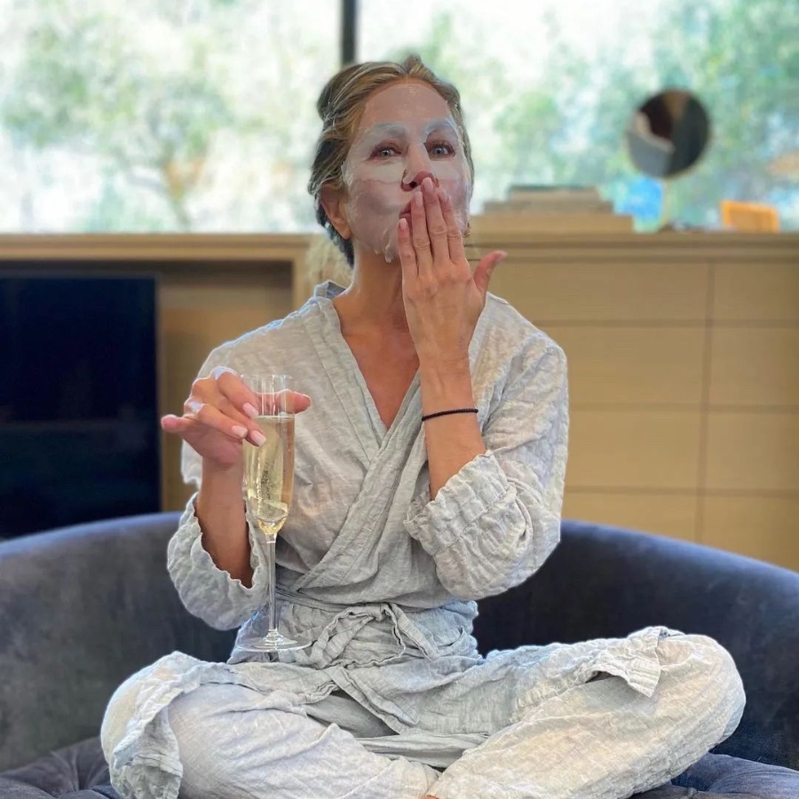 Jennifer Aniston wearing a sheet mask holding a glass of champagne