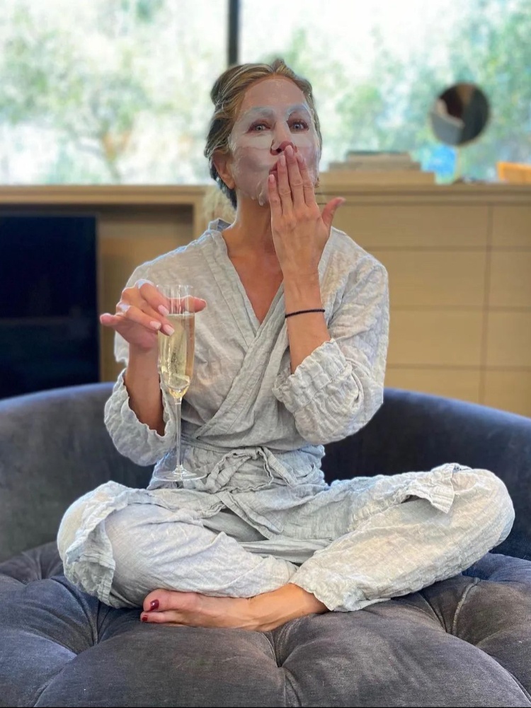 Jennifer Aniston wearing a sheet mask holding a glass of champagne